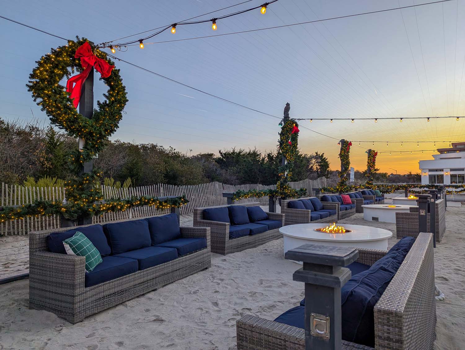 Beachside lounge area with gray wicker sofas with navy cushions, white fire pits, and string lights overhead. Large lit wreaths with red bows decorate the posts at sunset