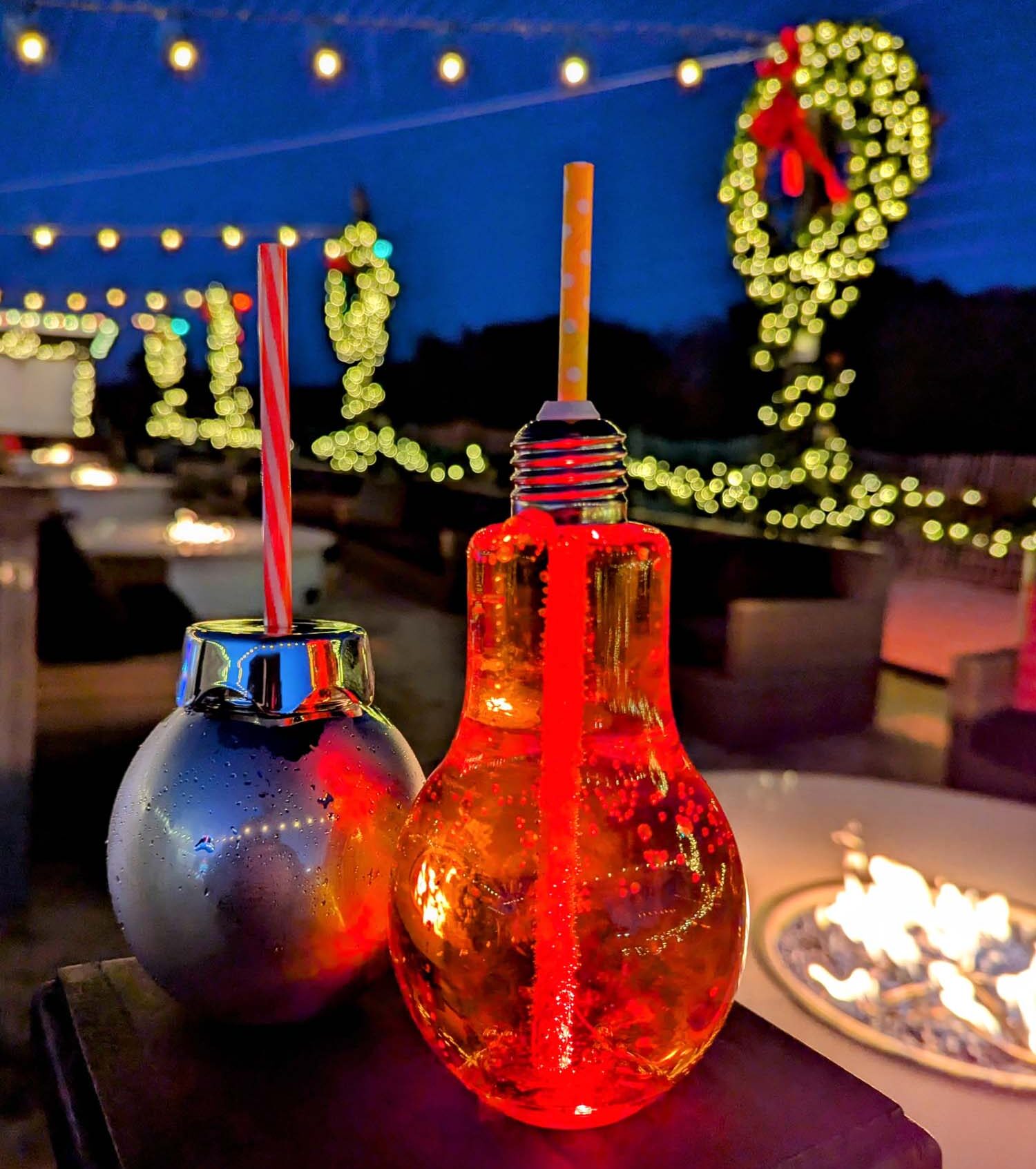 Close-up of two illuminated cocktail glasses shaped like Christmas ornaments - one blue and one red - with decorative straws, set against a backdrop of twinkling holiday lights