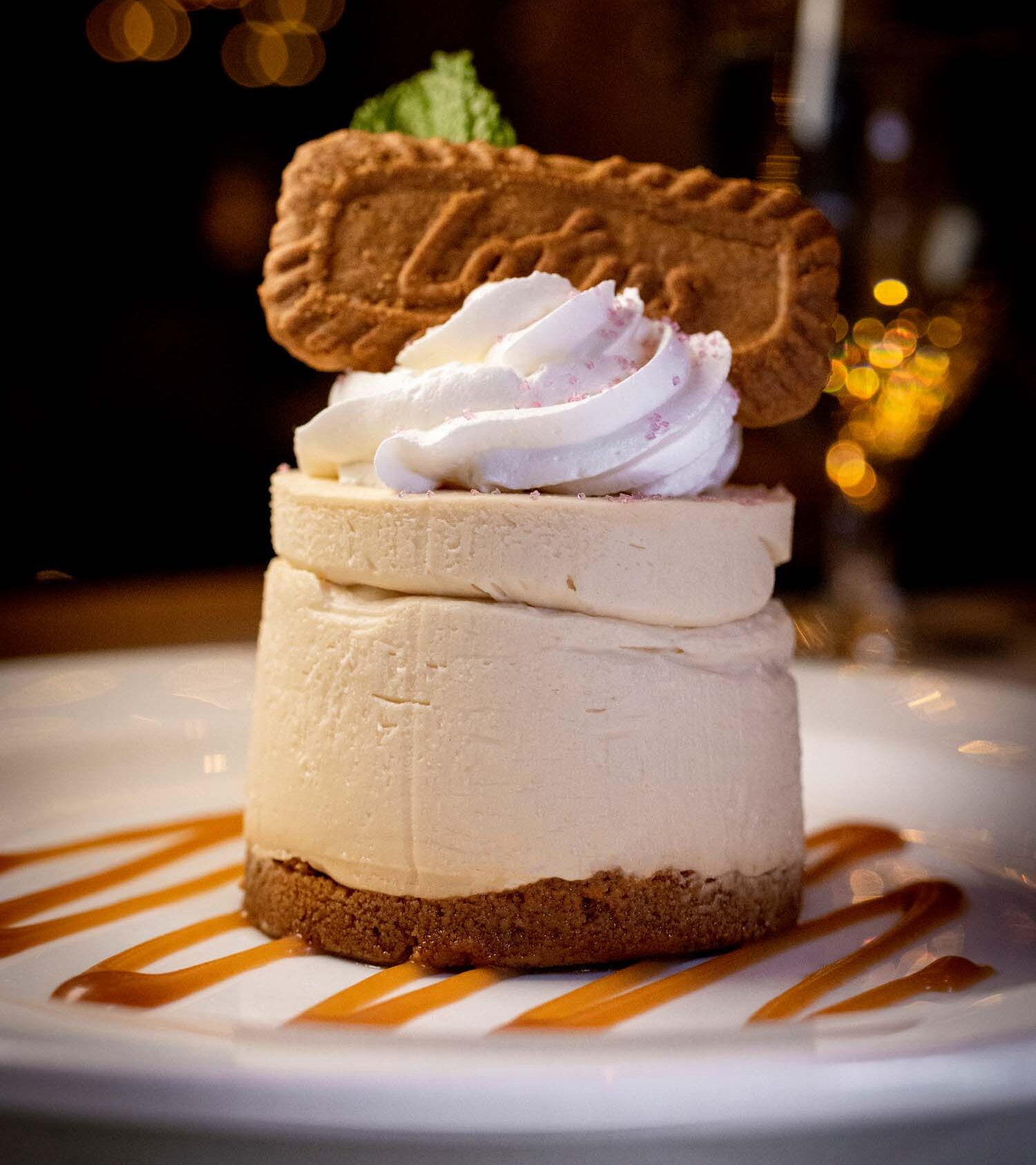 Towering New York-style cheesecake with Biscoff cookie crust, layered with whipped cream topping, garnished with a Biscoff cookie and mint leaf, served on caramel sauce stripes, photographed against warm bokeh holiday lights