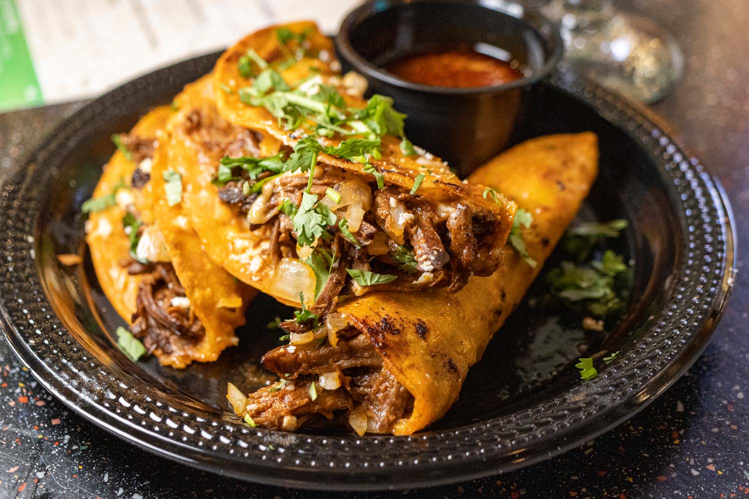 Crispy corn tortillas filled with braised beef birria, topped with melted cheese, fresh cilantro, and diced onions, served with a side of rich red consommé dipping sauce on a black textured plate