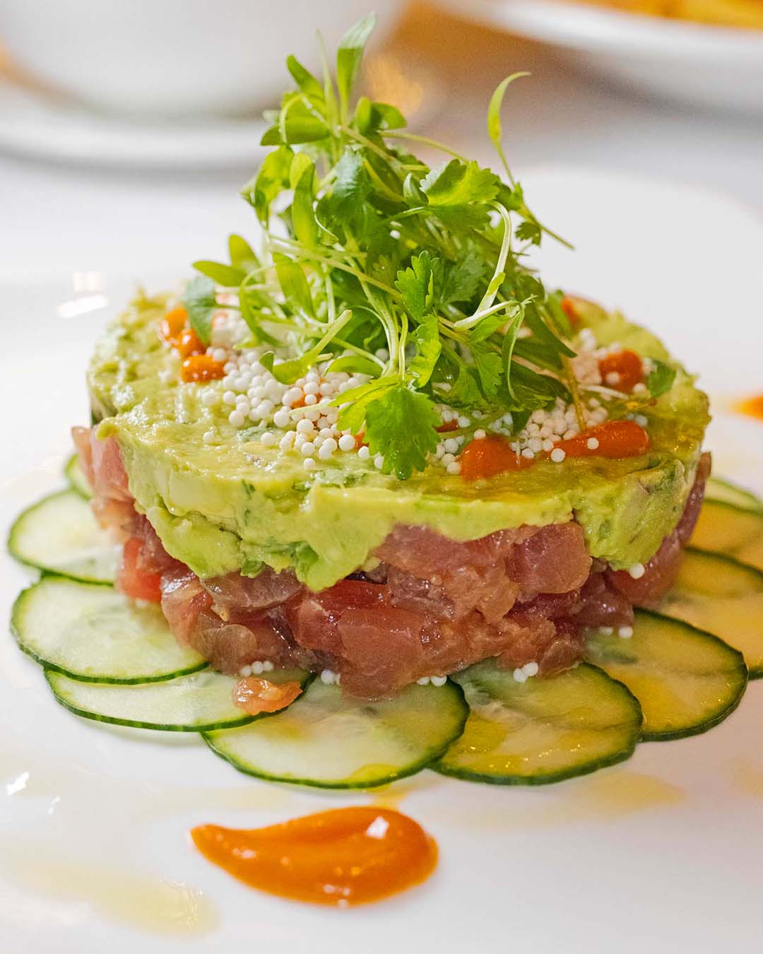 Layered tuna tartare and avocado tower garnished with micro greens, pearl caviar, and cucumber slices, served with citrus dots on a white plate