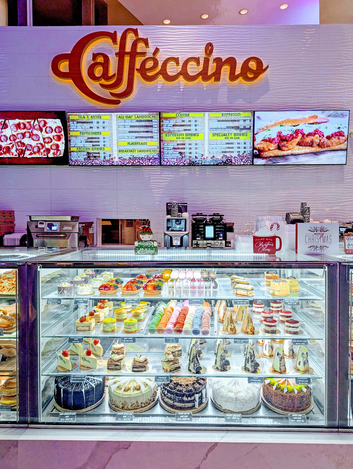 Caffecino cafe display case filled with colorful pastries, macarons, cakes and desserts, with illuminated menu boards and coffee machines above