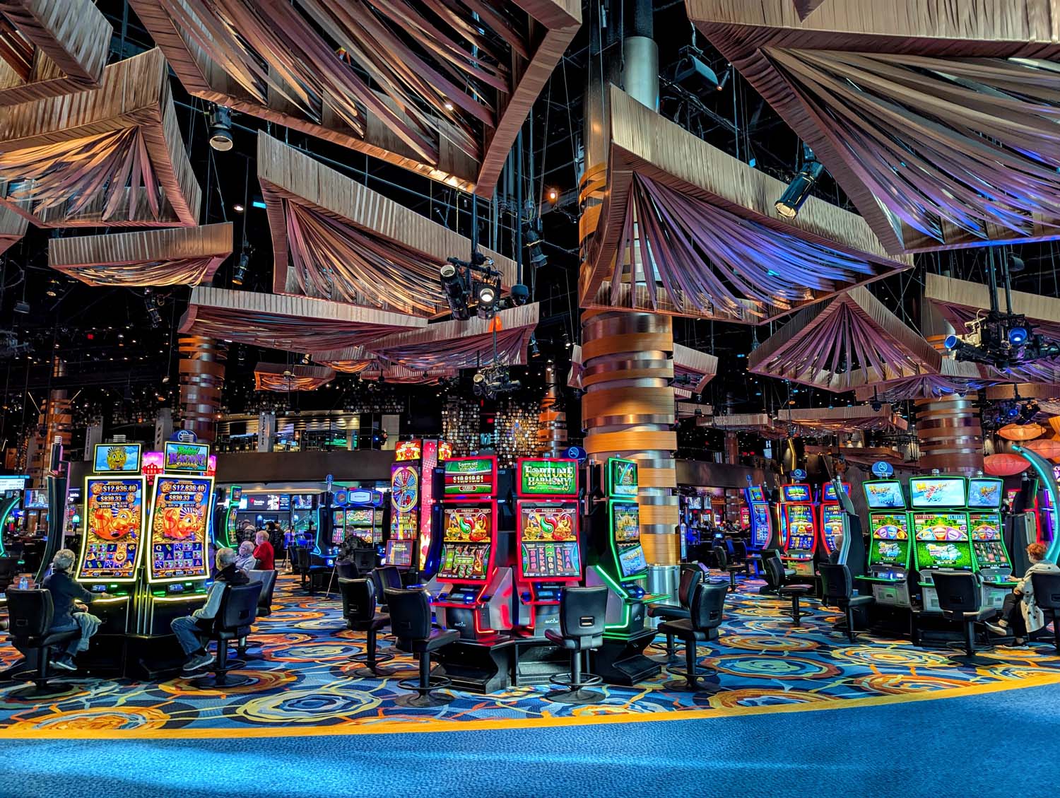 Modern casino gaming floor featuring illuminated slot machines beneath dramatic fabric ceiling installations, copper-toned columns, and vibrant LED displays with blue geometric patterned carpet, showcasing Ocean's contemporary design and gaming variety