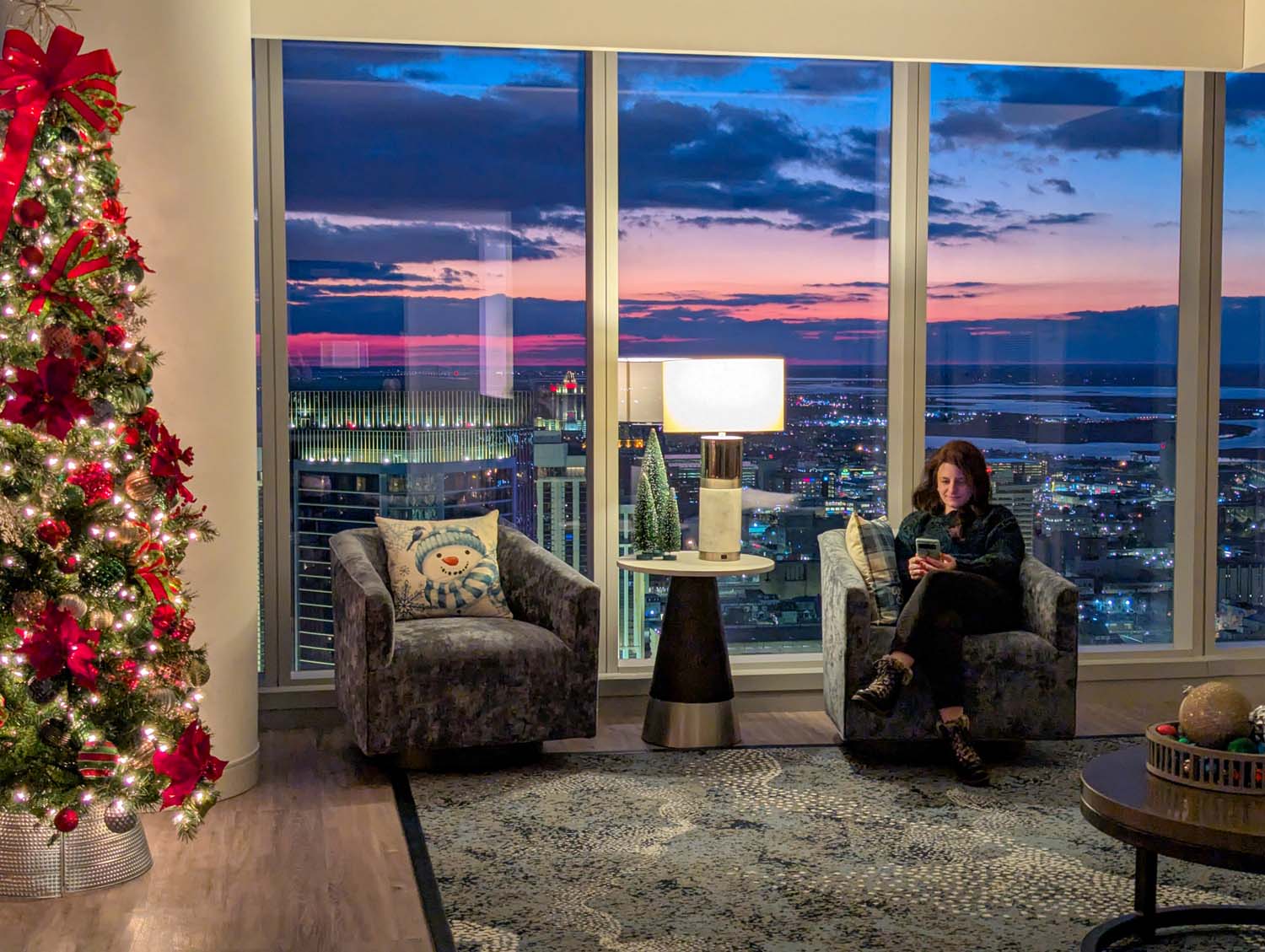 Corner suite living area with decorated Christmas tree, velvet chairs with snowman pillows, and floor-to-ceiling windows showcasing Atlantic City's dramatic sunset and city lights from the 36th floor of Ocean Casino Resort