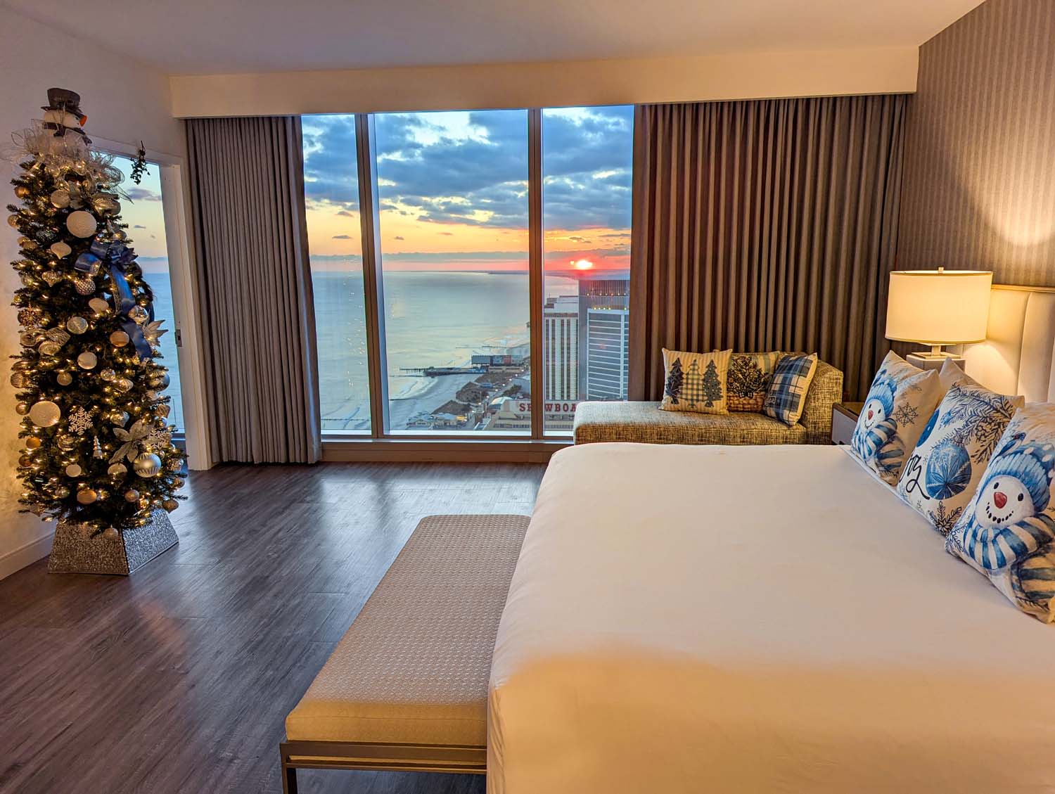 Hotel bedroom with dramatic sunset ocean view through floor-to-ceiling windows, decorated Christmas tree, white bedding with holiday pillows, and modern furnishings