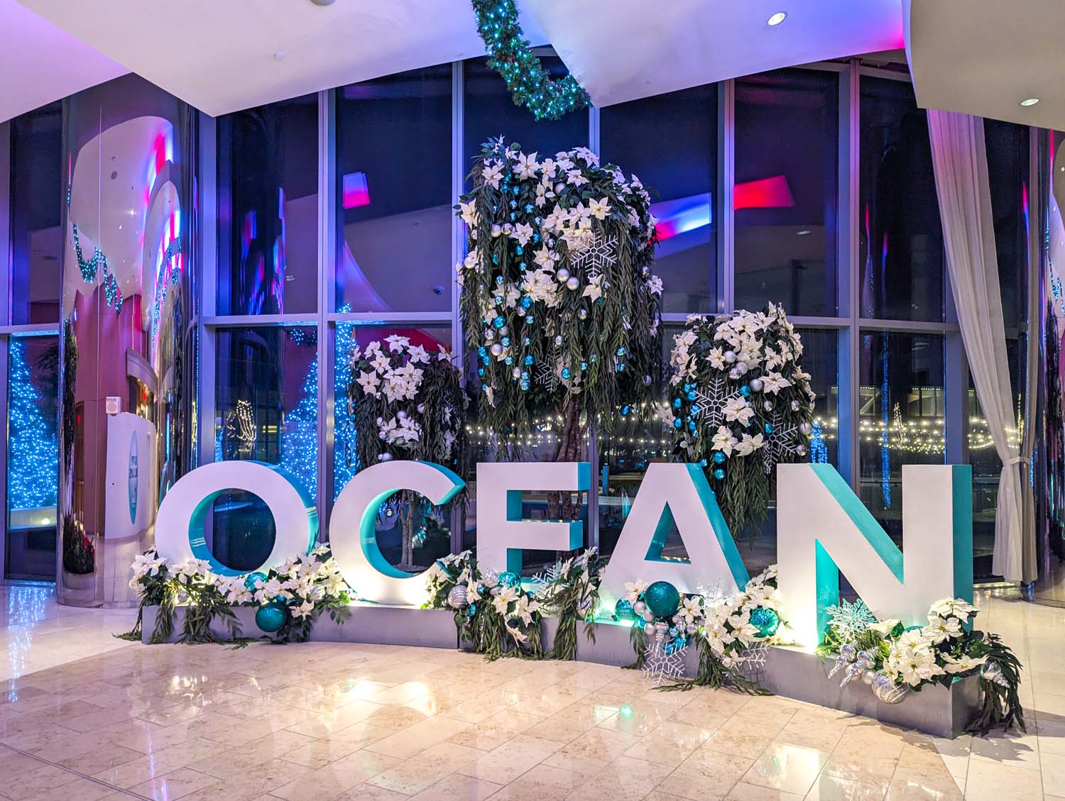 Large OCEAN letters display decorated with white flowers, turquoise lighting, snowflake accents, and holiday ornaments against a modern lobby backdrop