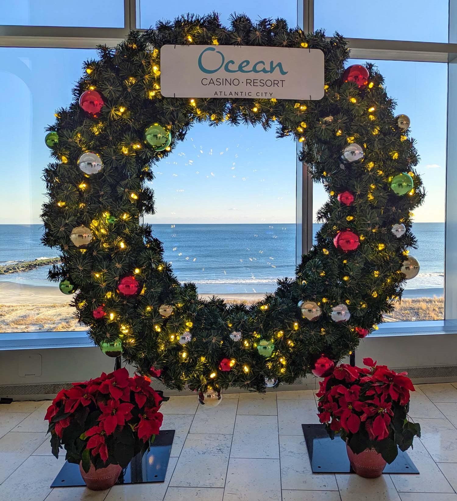 Oversized holiday wreath with Ocean Casino Resort signage framing a panoramic view of the Atlantic Ocean and beach, decorated with red, green, and gold ornaments, warm white LED lights, and flanked by bright red poinsettia plants on marble flooring, capturing both the festive atmosphere and coastal setting