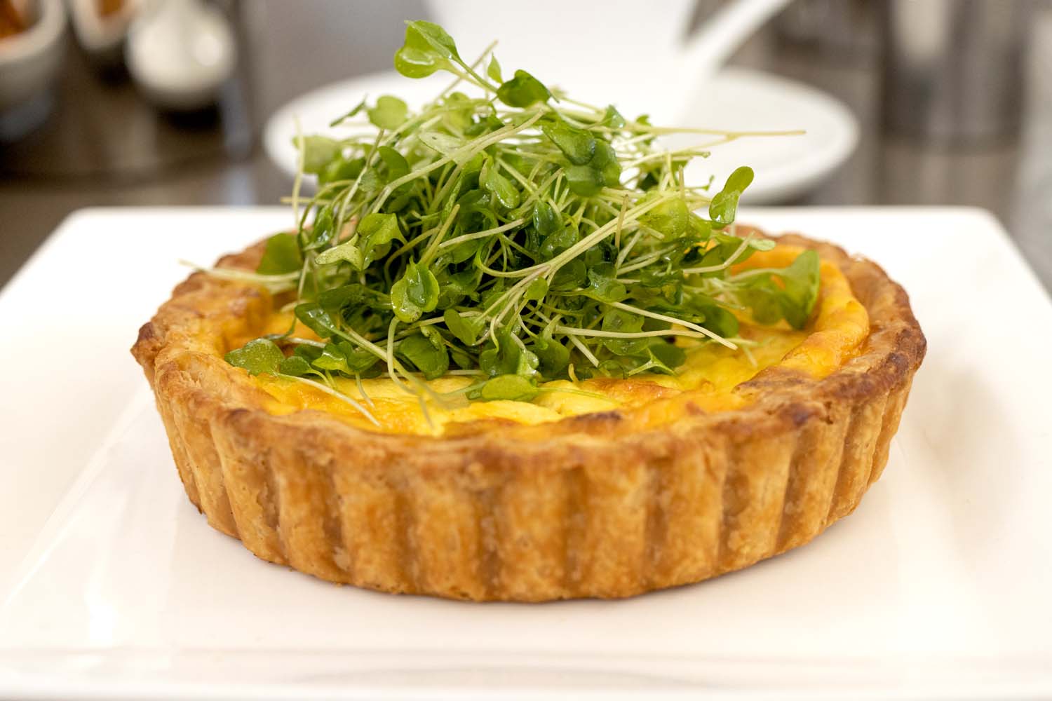 Individual quiche with golden brown flaky crust topped with fresh microgreens on a white plate