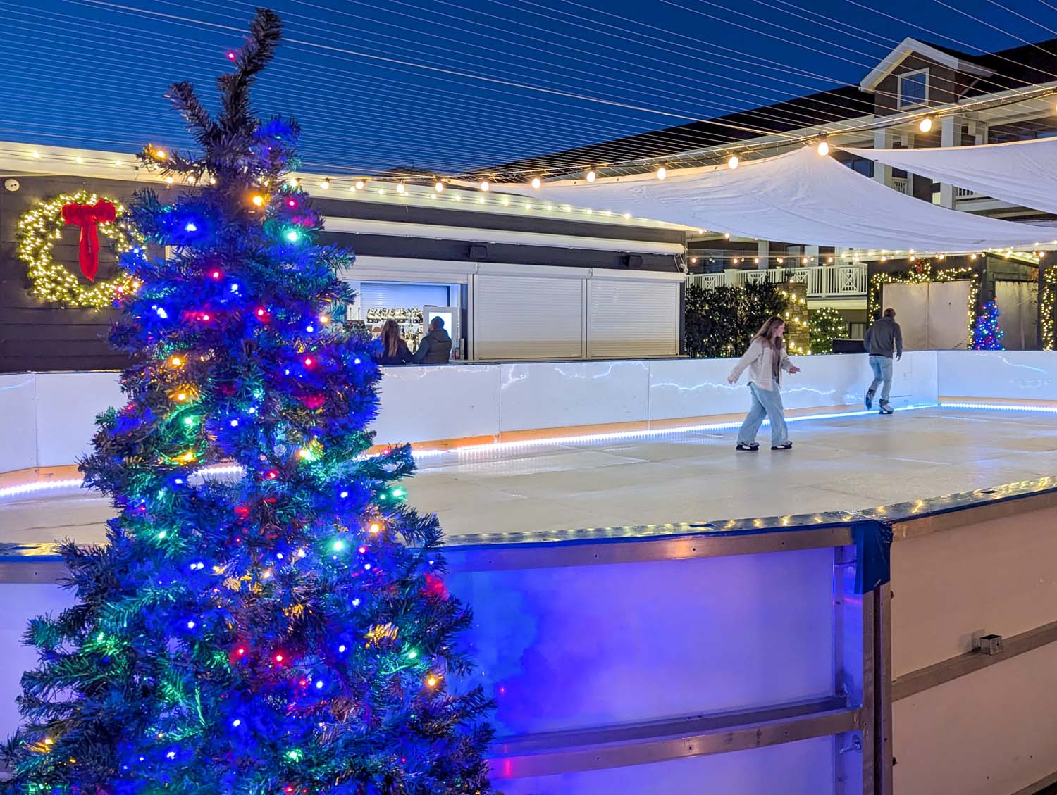 Illuminated synthetic ice skating rink at ICONA Avalon's Winter Wonderland, featuring a decorated Christmas tree with multicolored lights in the foreground, string lights overhead with white fabric canopies, and skaters enjoying the rink at dusk. A lit wreath with red bow hangs near the tree