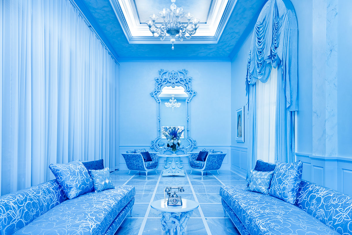 An elegant blue-toned salon with symmetrical layout featuring two damask-upholstered chaise lounges and ornate chairs arranged around marble-topped tables. A dramatic rococo mirror with carved details serves as focal point, crowned by a crystal chandelier visible in the coffered ceiling. The marble floor features geometric inlay patterns, and floor-length pleated curtains frame tall windows.