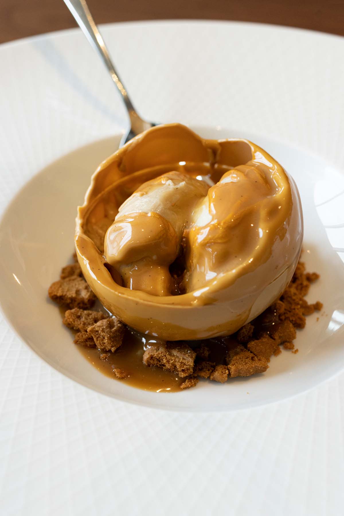 A spherical caramel-colored dessert on a white plate with chocolate crumbs, being drizzled with golden sauce that melts the outer shell