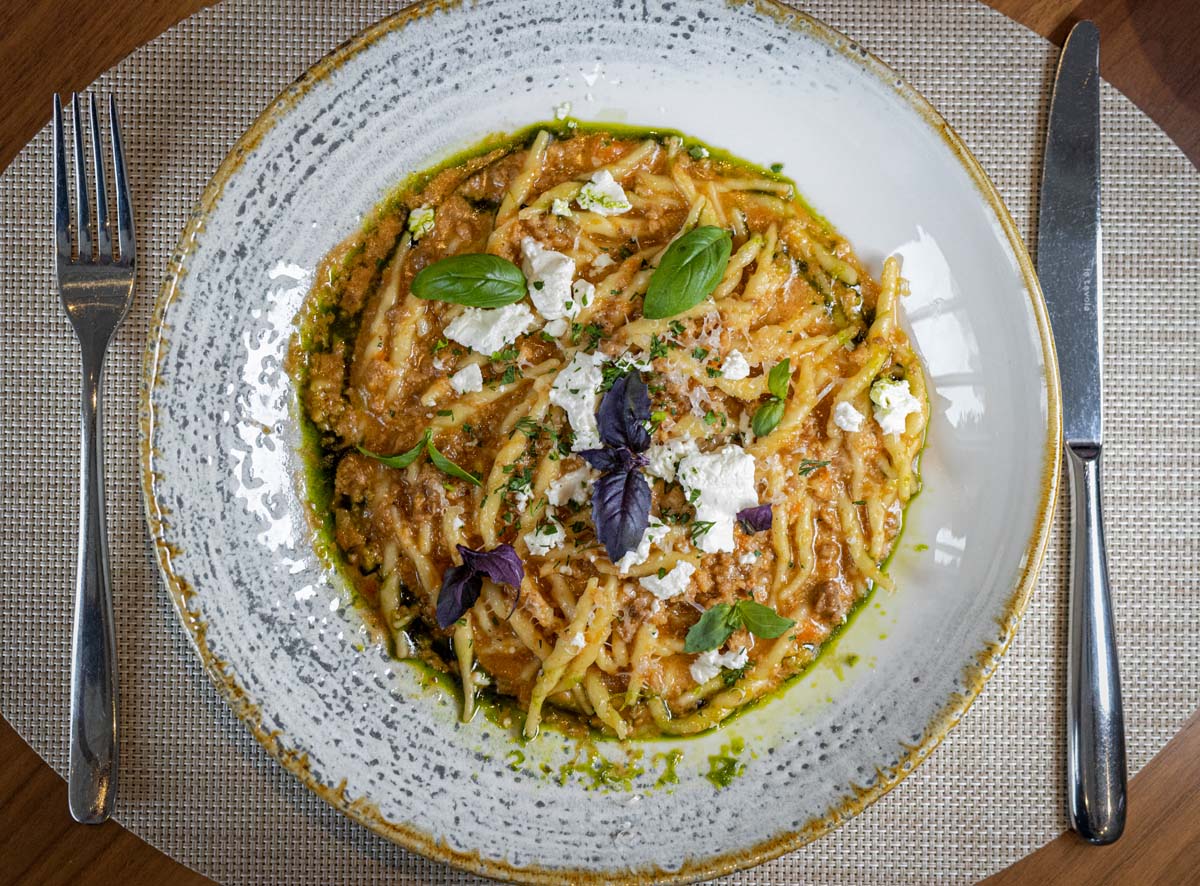 A plate of twisted pasta in a rich sauce, garnished with purple and green basil leaves and crumbled white cheese on a textured ceramic plate