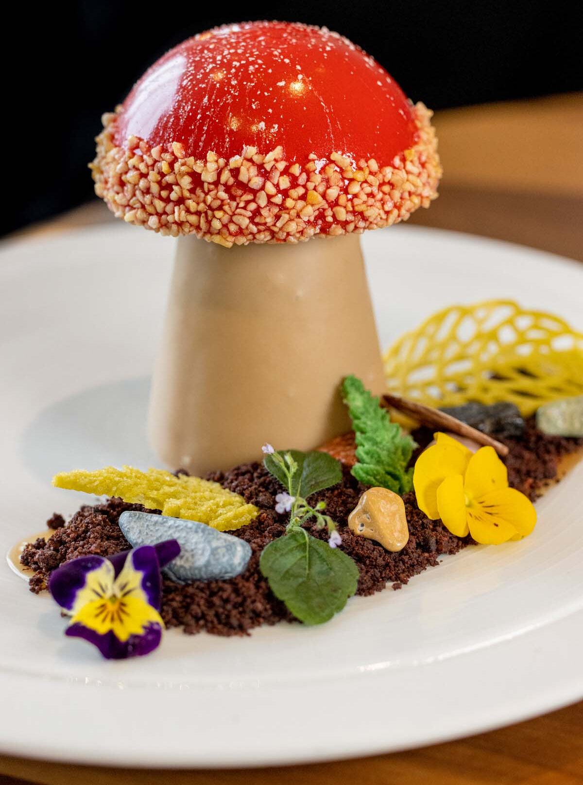 An artistic dessert designed to look like an amanita mushroom, featuring a glossy red dome with white speckles atop a beige stem. Set on a bed of chocolate 'soil' with decorative elements including yellow lace tuiles, edible flowers in purple and yellow, and fresh herbs. Served on a pristine white plate.