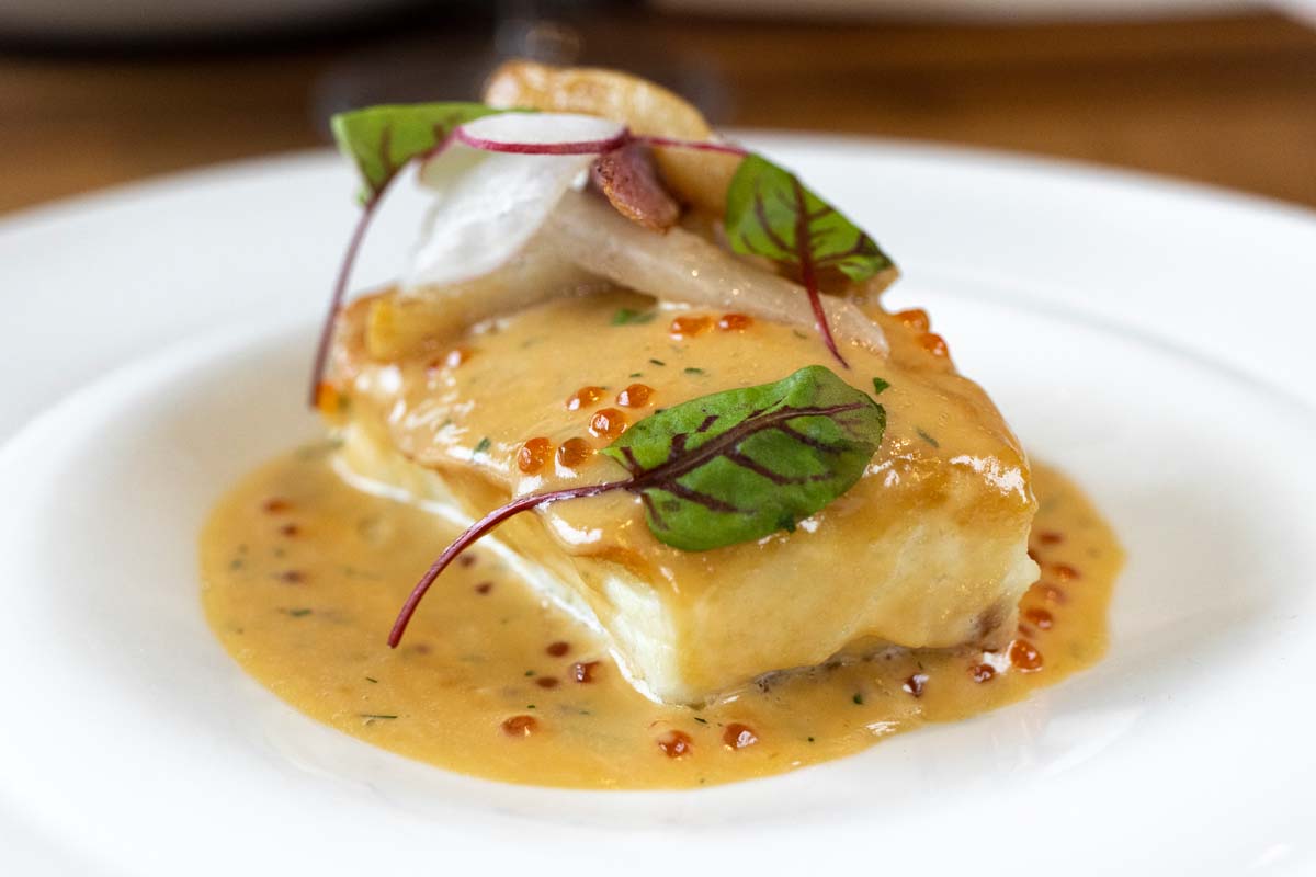 A perfectly seared piece of white halibut in a golden miso beurre blanc sauce, garnished with orange trout roe, thin slices of radish, and purple-veined sorrel leaves. The dish demonstrates refined plating on a white ceramic plate.