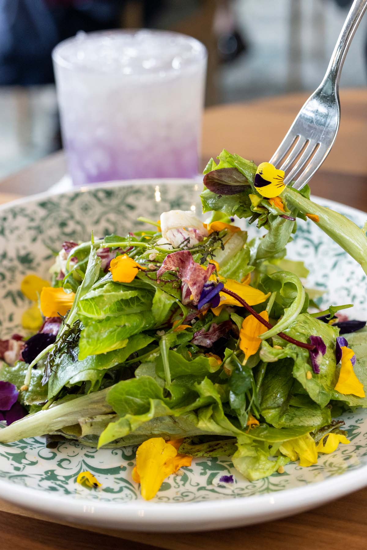 A fresh green salad with yellow and purple edible flowers, served on a decorative plate with a purple gradient cocktail in the background