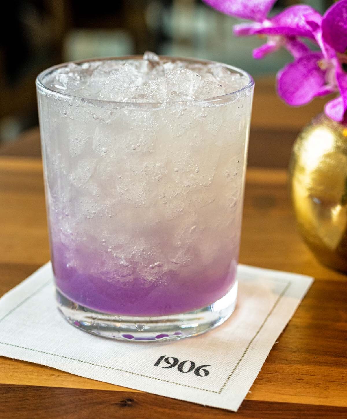 A tall glass containing a gradient cocktail that transitions from purple at the bottom to clear at the top, served on a napkin with "1906" branding