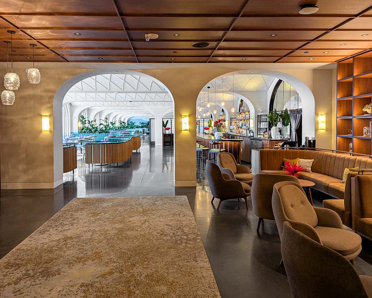 An elegant lounge space with arched entryways, wooden ceiling panels, and glass pendant lights. Caramel-colored velvet seating and built-in display cases showcase botanical specimens. The space flows through multiple arched doorways, creating a sense of depth and grandeur.