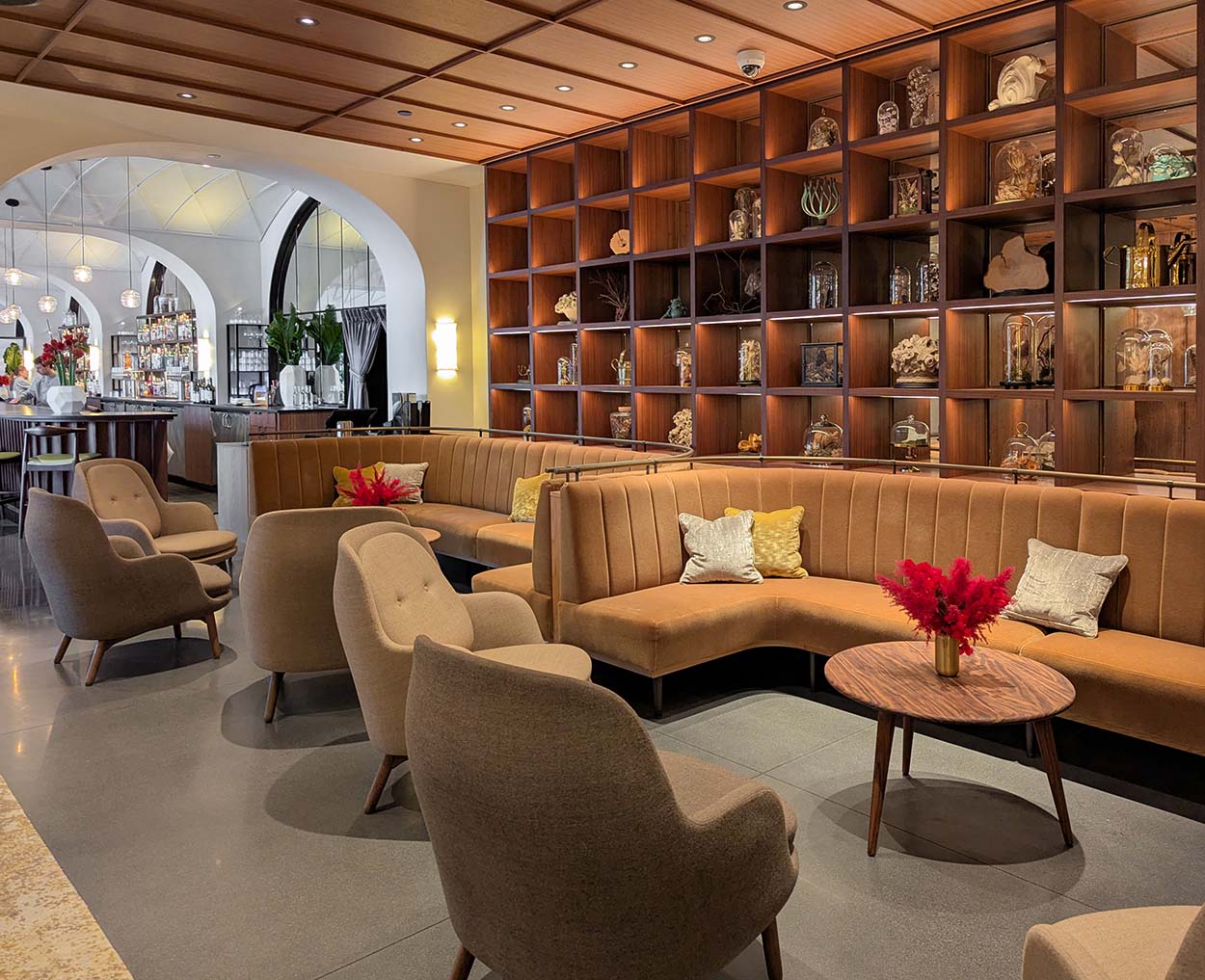 An elegant lounge area with tan curved banquettes, wooden accent tables, and wall-mounted display cases containing botanical specimens under glass