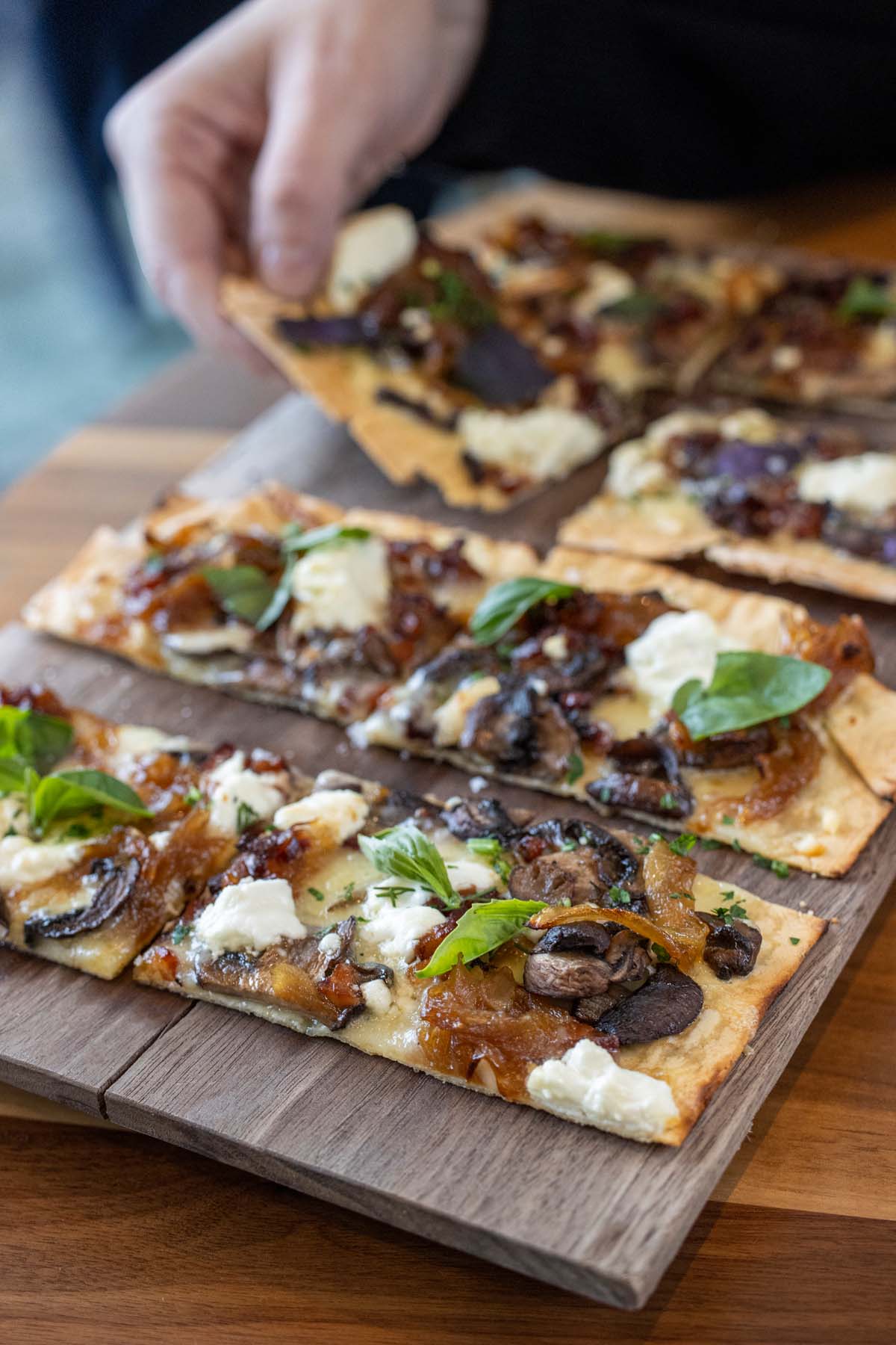 Flatbread cut into squares, topped with caramelized onions, mushrooms, white cheese, and fresh herbs, served on a wooden board