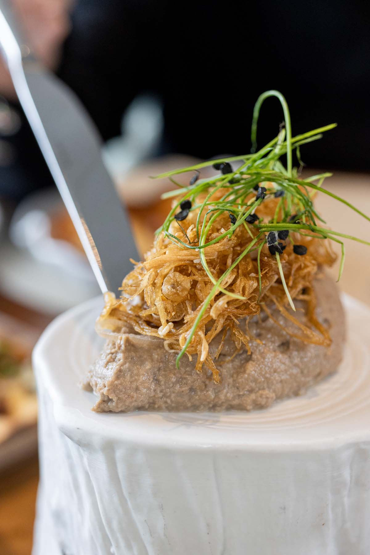 A grey-colored pâté topped with crispy golden mushrooms and fresh green herbs, served on a white plate