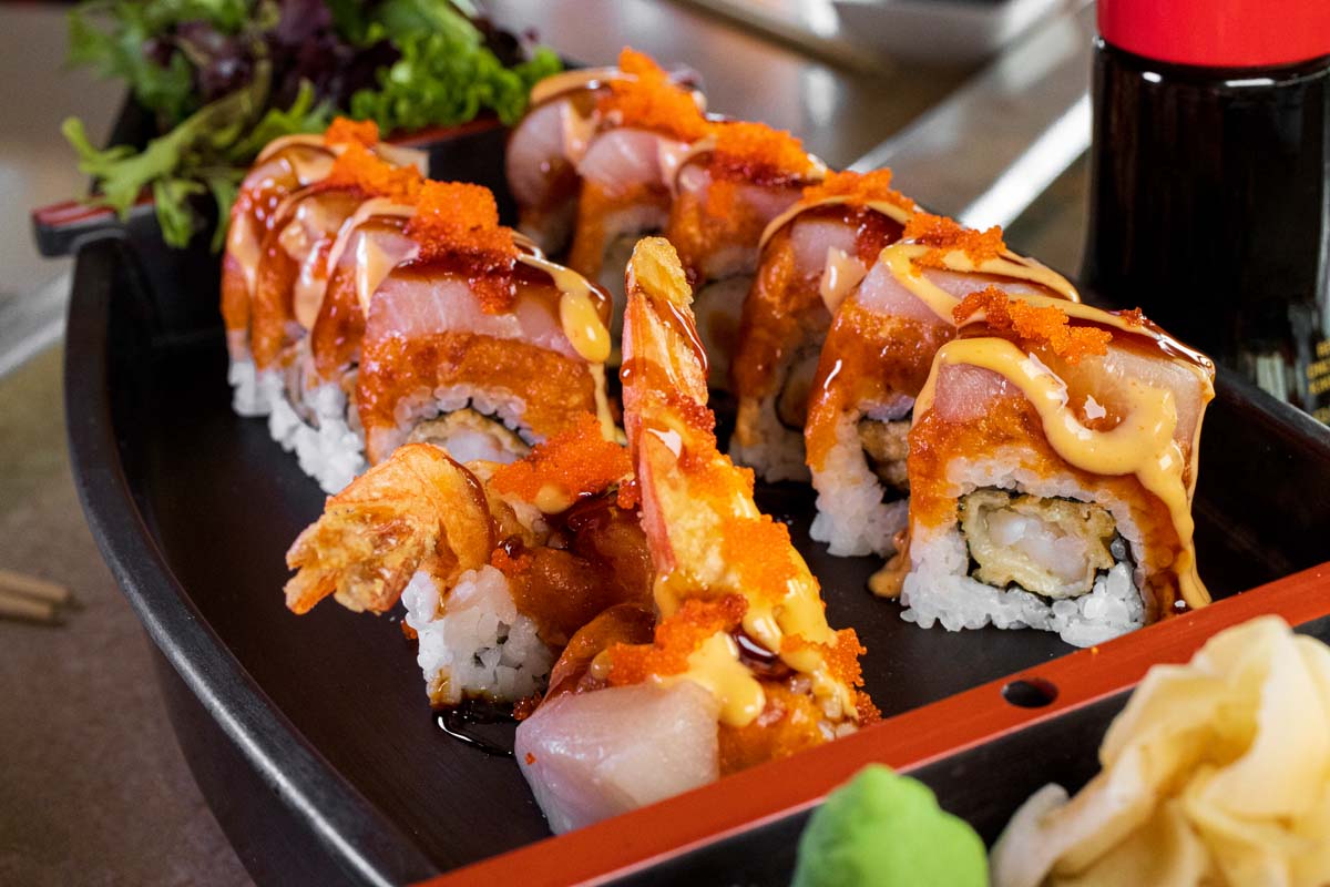 Artfully presented sushi roll featuring tempura shrimp, yellowtail, and spicy tuna, topped with tobiko, drizzled with eel sauce and spicy mayo, served on a black plate with fresh greens