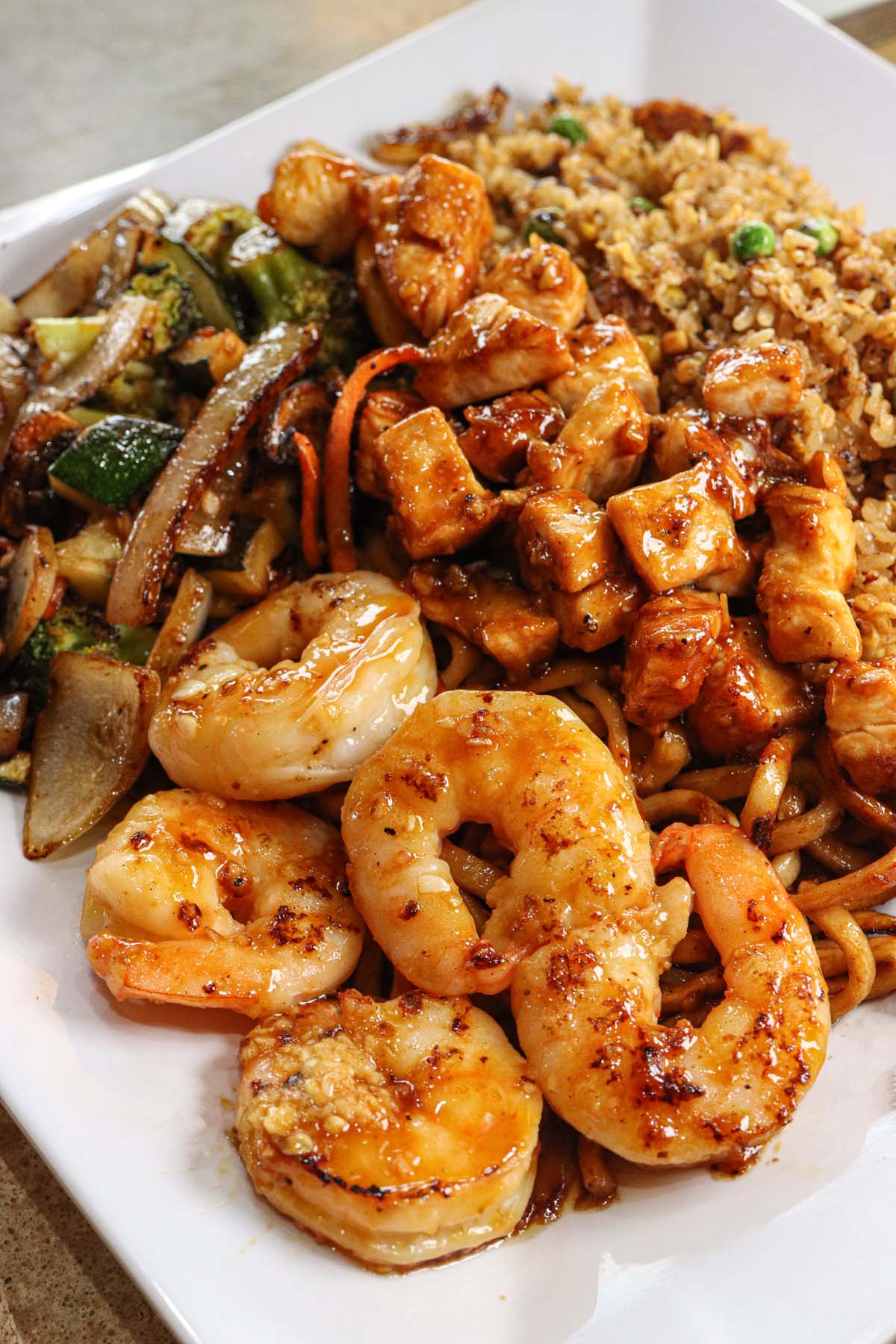 Close-up of hibachi-grilled jumbo shrimp and chicken with stir-fried vegetables, including onions, zucchini, and broccoli, served with fried rice, all glistening with teriyaki sauce