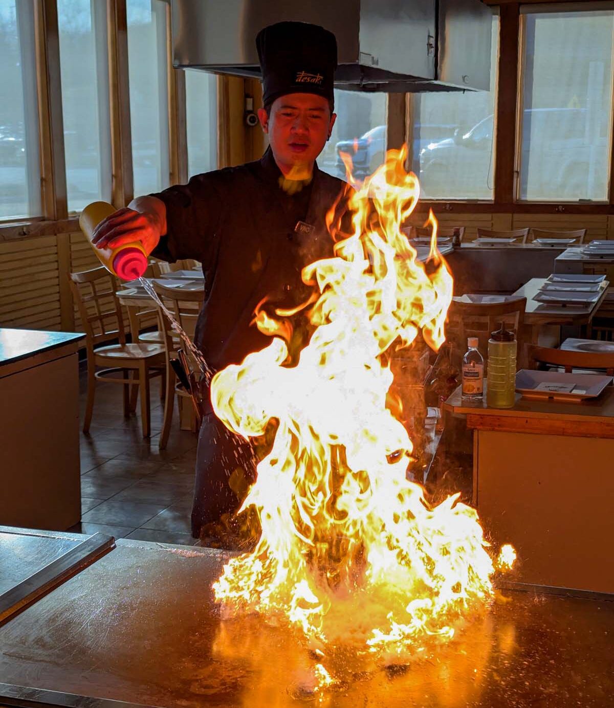 Hibachi chef in traditional uniform and hat creating dramatic flames on the grill, illuminating the dining room with the signature Desaki fire show