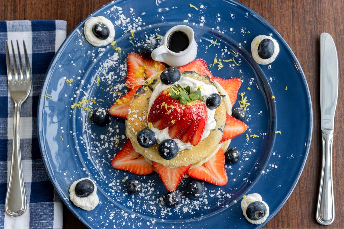 A stack of fluffy pancakes served on a navy blue plate, garnished with fresh strawberries, blueberries, powdered sugar, and dollops of whipped cream decorated with fresh blueberries. A small pitcher of maple syrup sits alongside on the plate. The presentation includes decorative dots of whipped cream around the plate's rim, creating an elegant breakfast presentation.