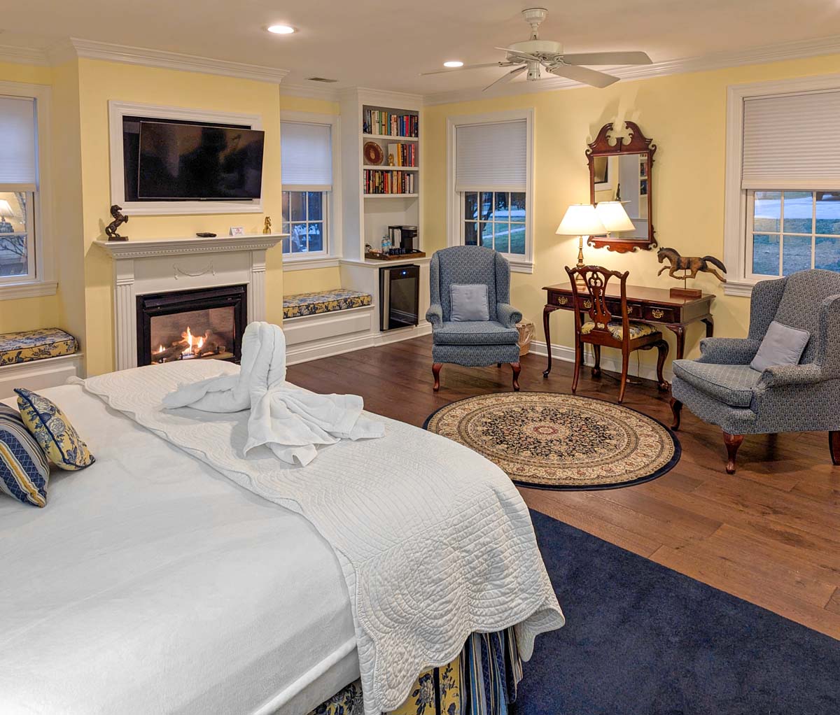 A spacious guest room with buttery yellow walls and white trim, featuring a king-size bed with white linens, built-in window seats with floral cushions, a large flat-screen TV mounted above a white fireplace mantel, blue wingback chairs, and antique wooden furniture. Multiple windows provide natural light and garden views.