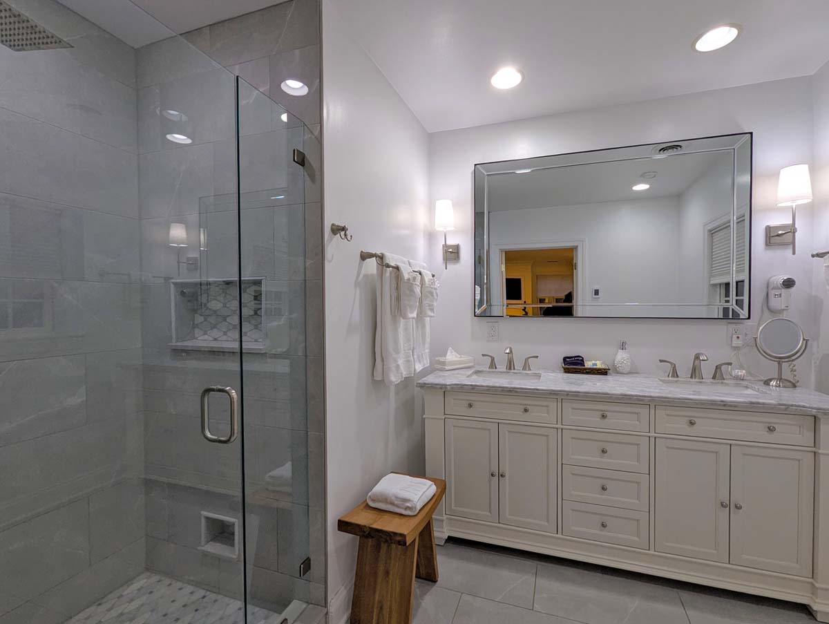 A modern bathroom with heated marble floors, featuring a large glass-enclosed shower with gray tile, a double vanity with white cabinetry and marble countertop, brushed nickel fixtures, a large framed mirror, and elegant sconce lighting. White towels and high-end amenities complete the spa-like setting.