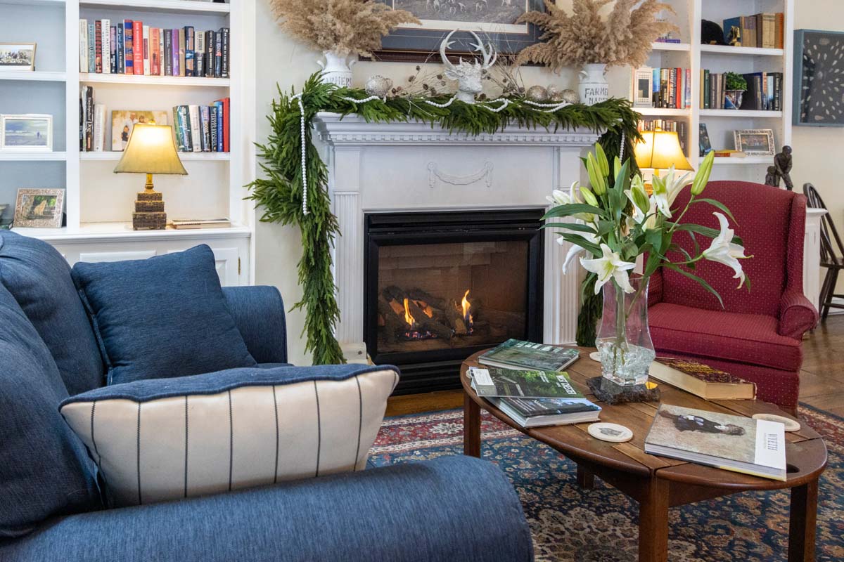 A welcoming sitting room with built-in white bookshelves, a traditional fireplace decorated with winter greenery and pampas grass, a navy blue sofa, burgundy wingback chair, and oriental rug. Fresh white lilies in a vase and table lamps create a warm atmosphere.