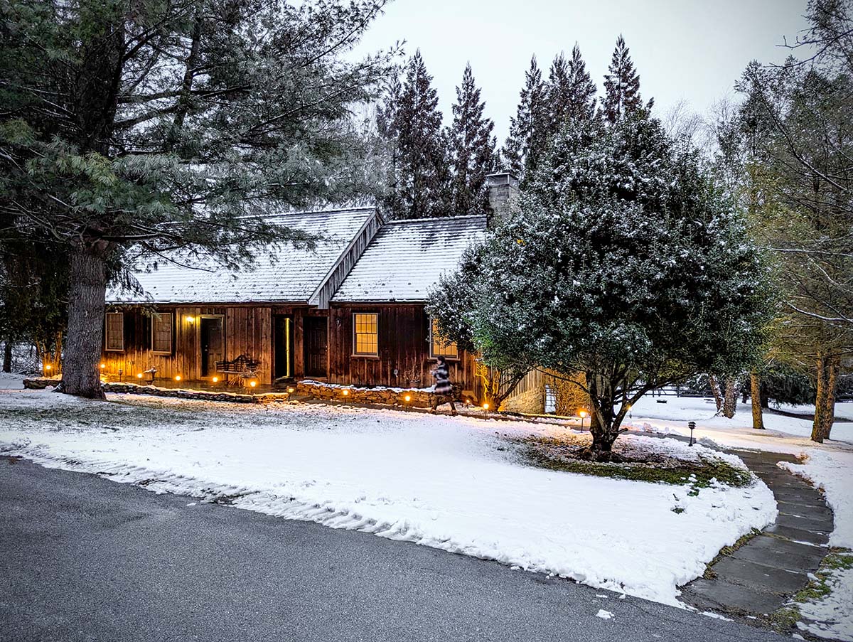 A rustic wooden barn with warm lighting glowing from within, set against a snowy evening scene. The barn features cedar shake siding, stone foundation, and pathway lights creating an inviting atmosphere. Snow-covered evergreens and pathway lights add to the cozy winter ambiance.