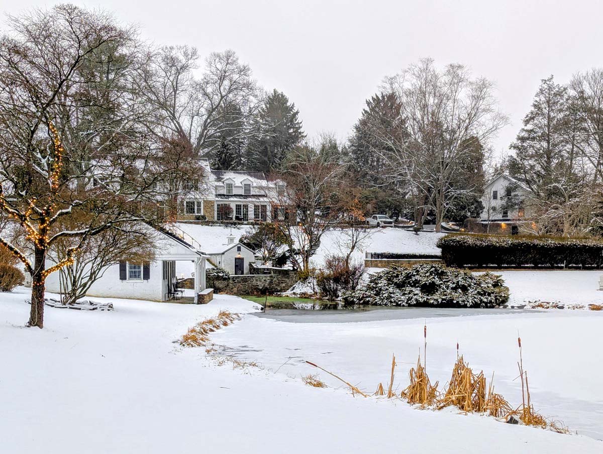 A panoramic view of the property showing a white colonial-style main house with dormers, a smaller outbuilding, and manicured grounds covered in snow. A tree decorated with white lights stands in the foreground, and dried ornamental grasses peek through the snow near a frozen pond.