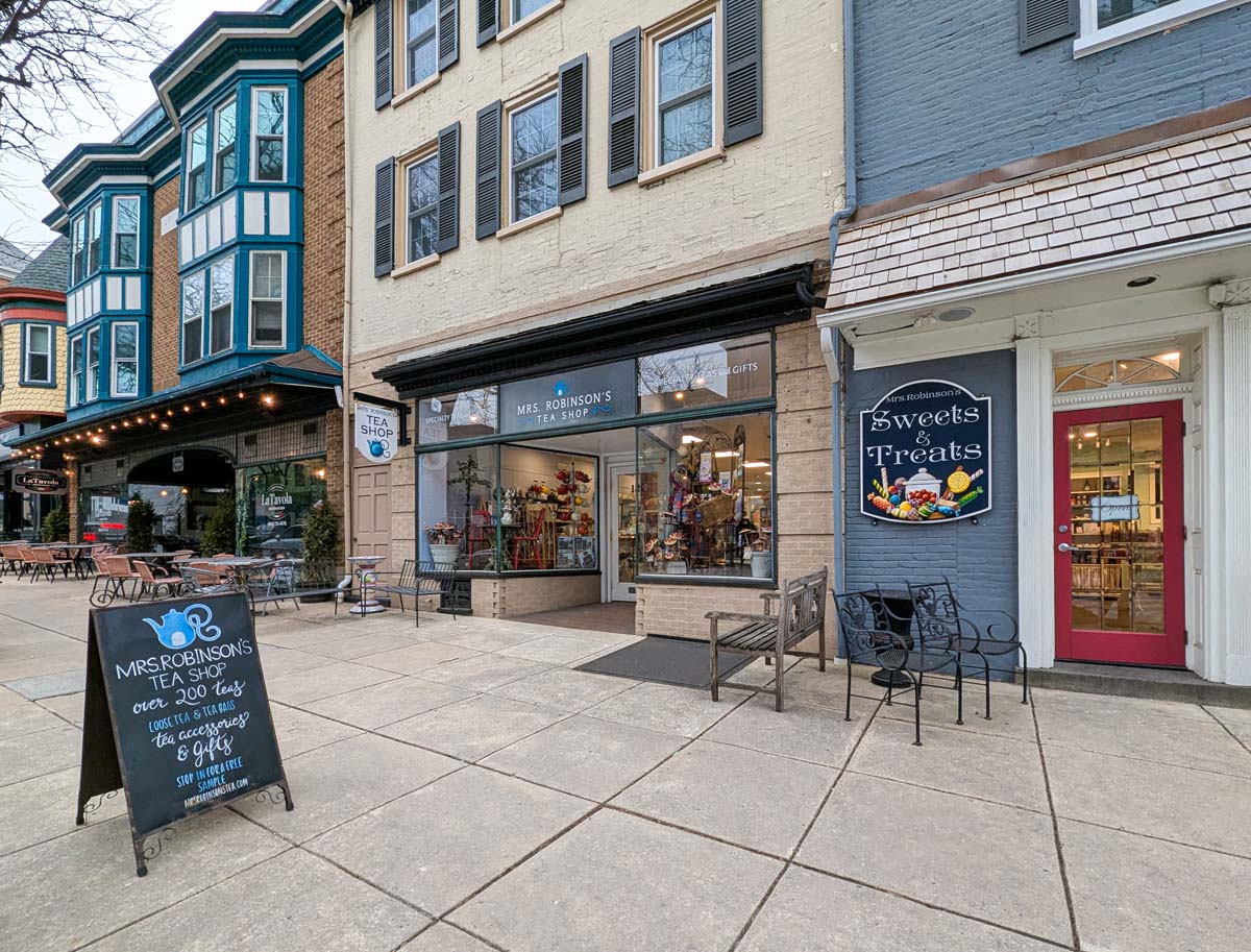 Historic brick storefront with cream-colored facade and bay windows painted turquoise. The shop features two connected businesses: Mrs. Robinson's Tea Shop with its signature blue teapot logo and Sweets & Treats with a red door. Wrought iron benches and a sidewalk chalkboard sign advertising over 200 teas complete the welcoming entrance.