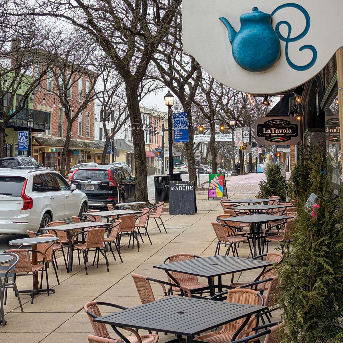 Charming streetscape of downtown Kennett Square with bare winter trees, vintage-style lampposts with town banners, and outdoor dining furniture from La Tavola restaurant. Historic brick buildings and locally-owned shops line the street, while the blue teapot sign from Mrs. Robinson's Tea Shop is visible in the foreground.