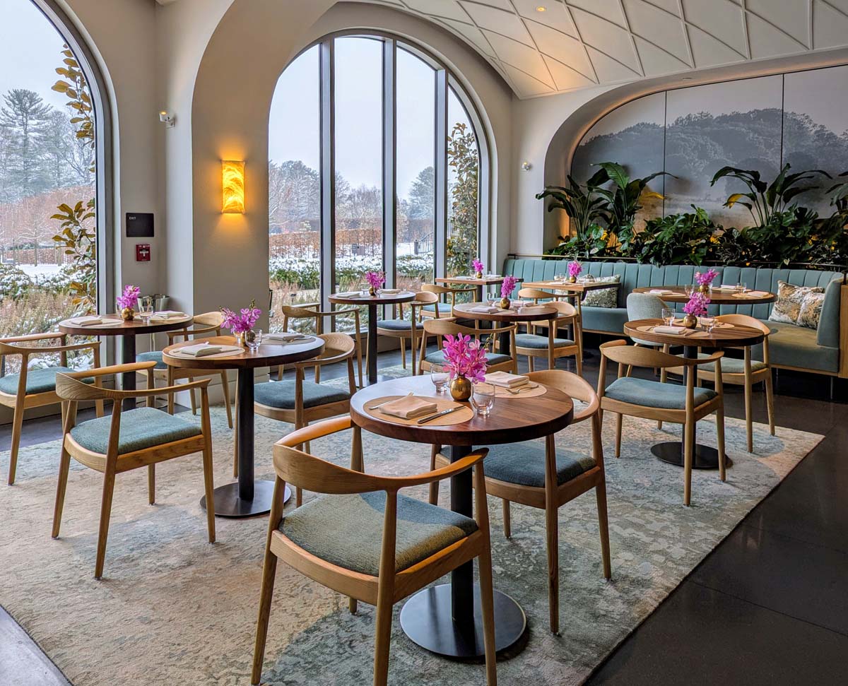 An elegant restaurant interior with soaring arched windows, wooden tables with mid-century modern chairs upholstered in sage green fabric. Pink orchid centerpieces adorn each table. A built-in banquette with sea-green upholstery runs along one wall, backed by lush indoor plants. Warm amber wall sconces provide atmospheric lighting against white walls. The windows frame a snowy winter landscape beyond.