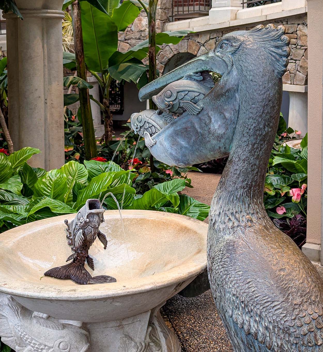 A decorative bronze fountain featuring a large pelican-like bird with fish spouts, creating streams of water into a stone basin. The fountain is set against a backdrop of tropical foliage and stone architecture with bananas leaves overhead.