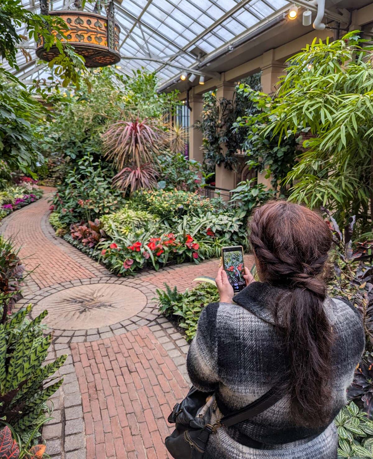 A visitor photographing a curved brick pathway with their phone, surrounded by lush tropical plantings and illuminated by decorative overhead lanterns. The path features intricate brick patterns and is bordered by colorful foliage and flowers.