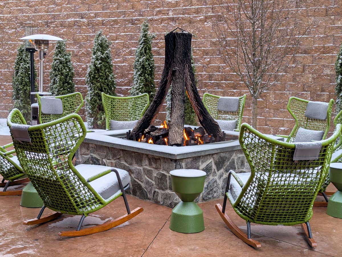 Intimate outdoor seating area with modern lime-green woven rocking chairs arranged around a stone firepit with active flames, decorated with snow-covered evergreens against a terraced stone wall