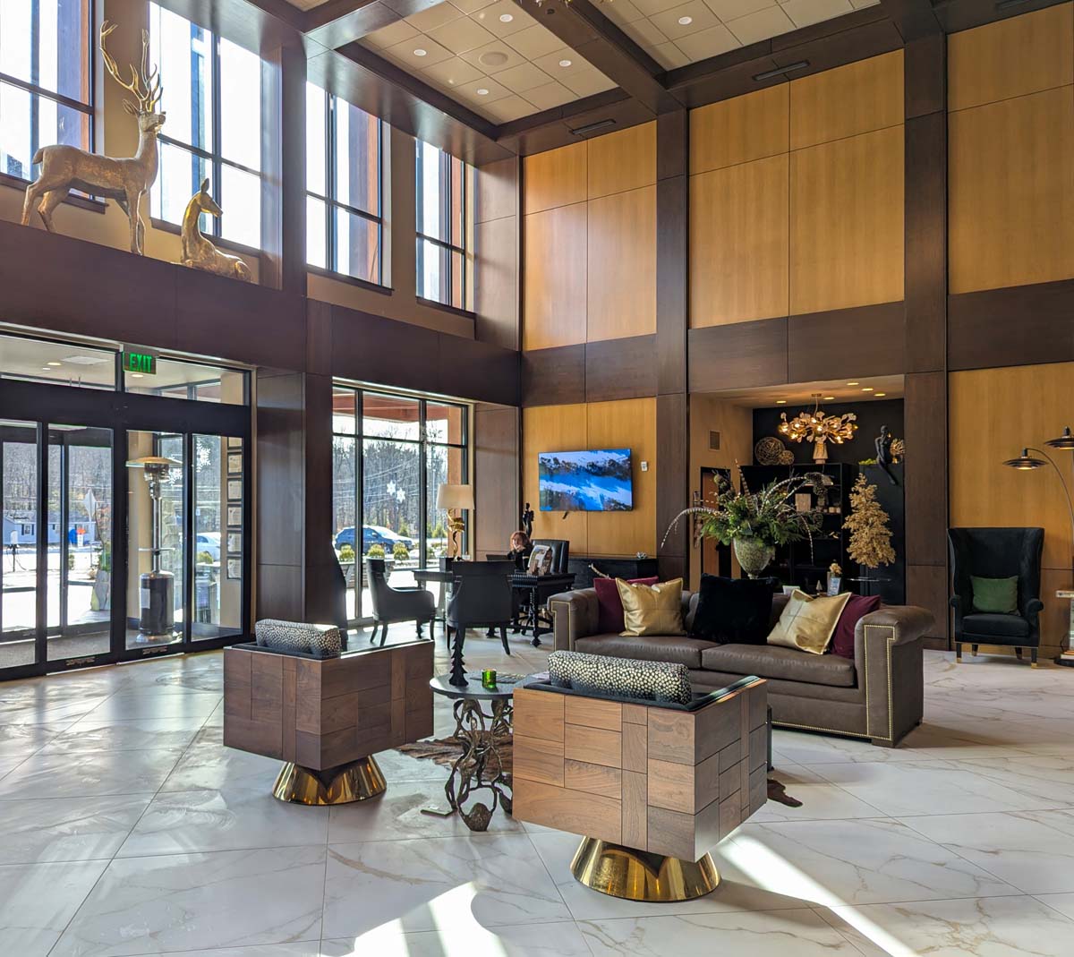 Double-height hotel lobby with wood and gold accents, marble floors, modern furniture, and decorative deer sculptures in window