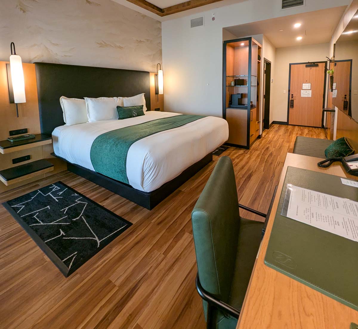 Luxurious hotel room featuring a king bed with crisp white linens and forest green accents, hardwood floors, modern wall sconces, custom area rug, and built-in workstation with green leather chair