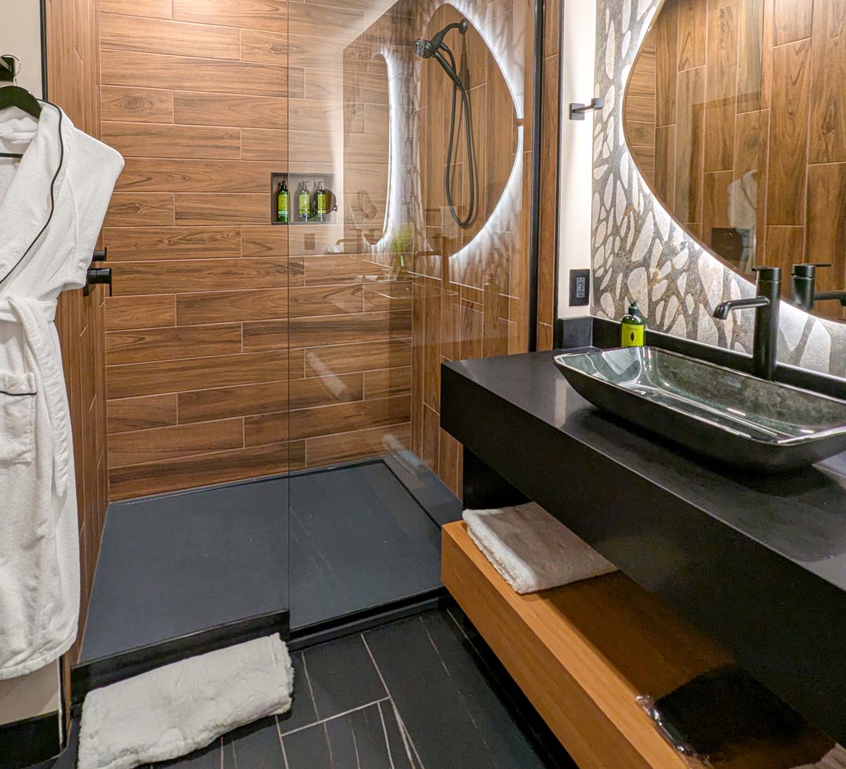 Modern bathroom with wood-look tile walls, illuminated circular mirror, vessel sink on black vanity, rain shower with glass door, built-in toiletry niche, and plush white robes