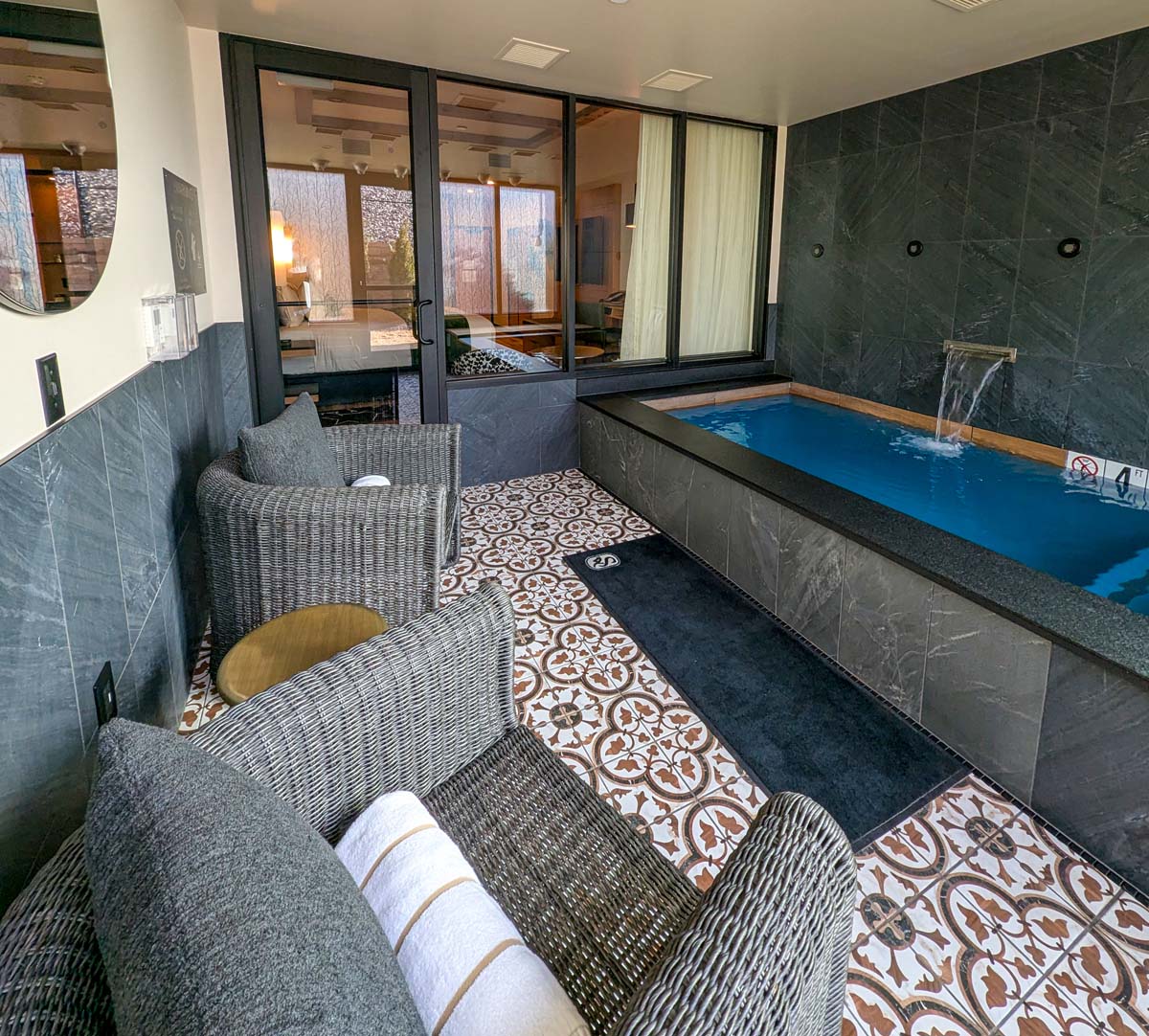 Luxurious private pool room with patterned tile floor, grey stone walls, waterfall feature, and comfortable wicker lounge chairs