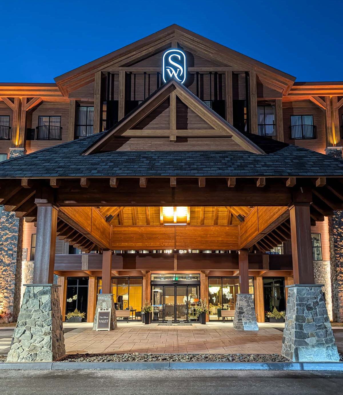 Dramatic nighttime view of the hotel entrance featuring illuminated timber-frame porte-cochère, glowing Swiftwater logo, stone pillars, and warm wood architectural details against deep blue sky