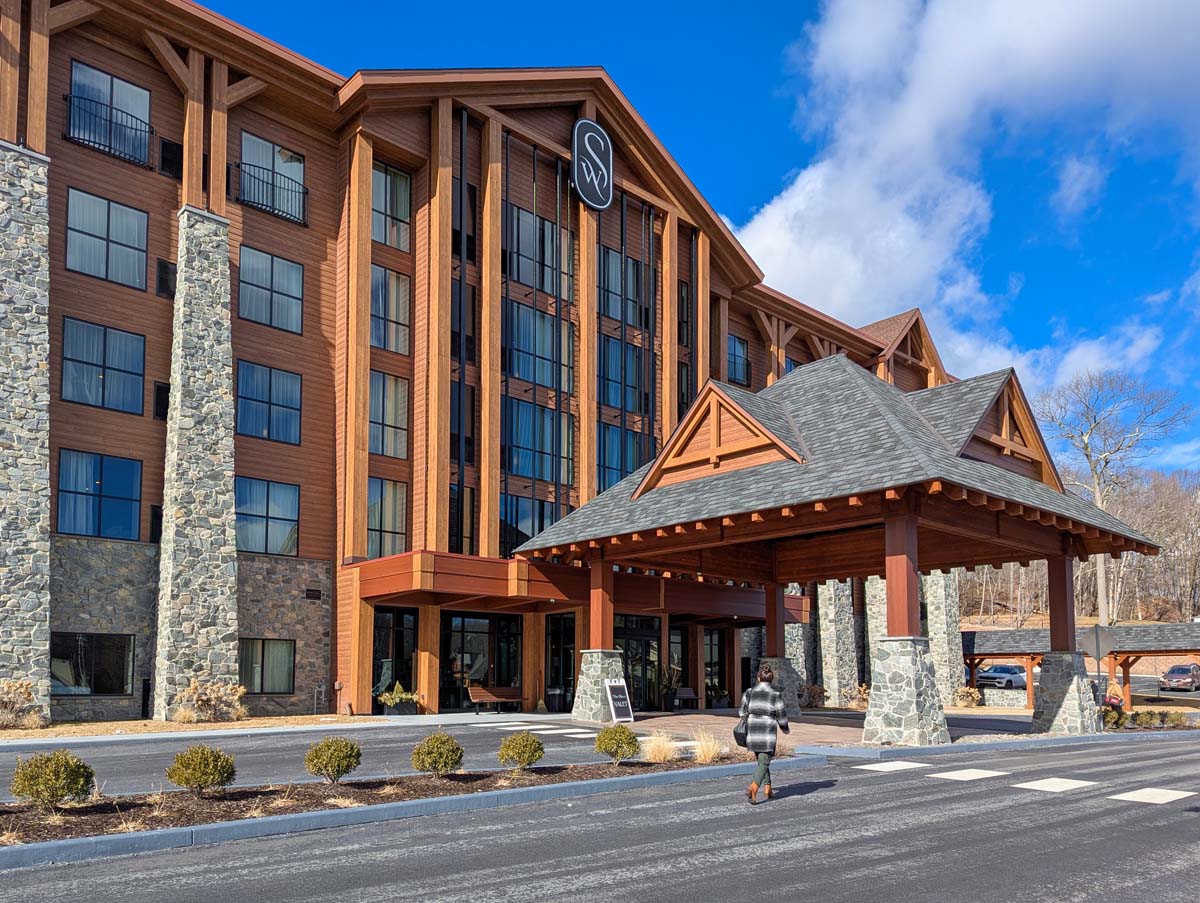 Exterior of modern mountain hotel featuring stone pillars, warm wood siding, dramatic floor-to-ceiling windows, covered porte-cochère entrance with timber frame details, and branded signage