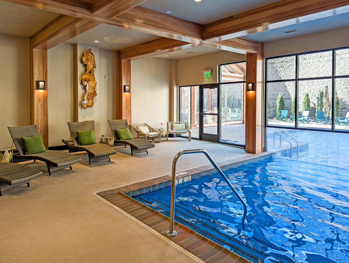 Indoor pool area with floor-to-ceiling windows, exposed wooden beams, natural wood accent wall with artistic driftwood piece, grey woven chaise lounges with green pillows, and blue pool with brick border