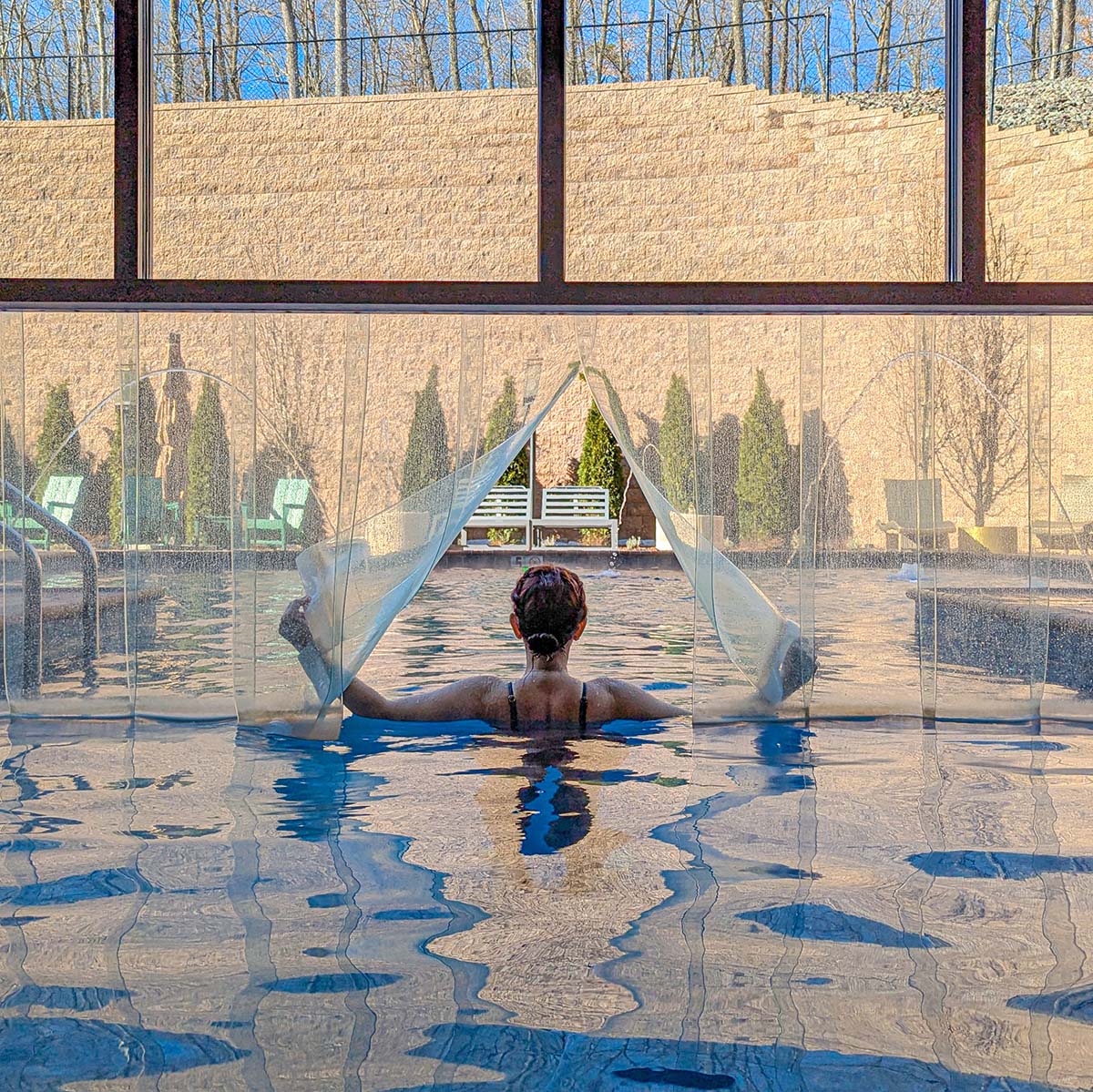 Person enjoying the heated indoor/outdoor pool at The Swiftwater Hotel, holding the water features while overlooking snow-dusted evergreens and stone wall through floor-to-ceiling windows