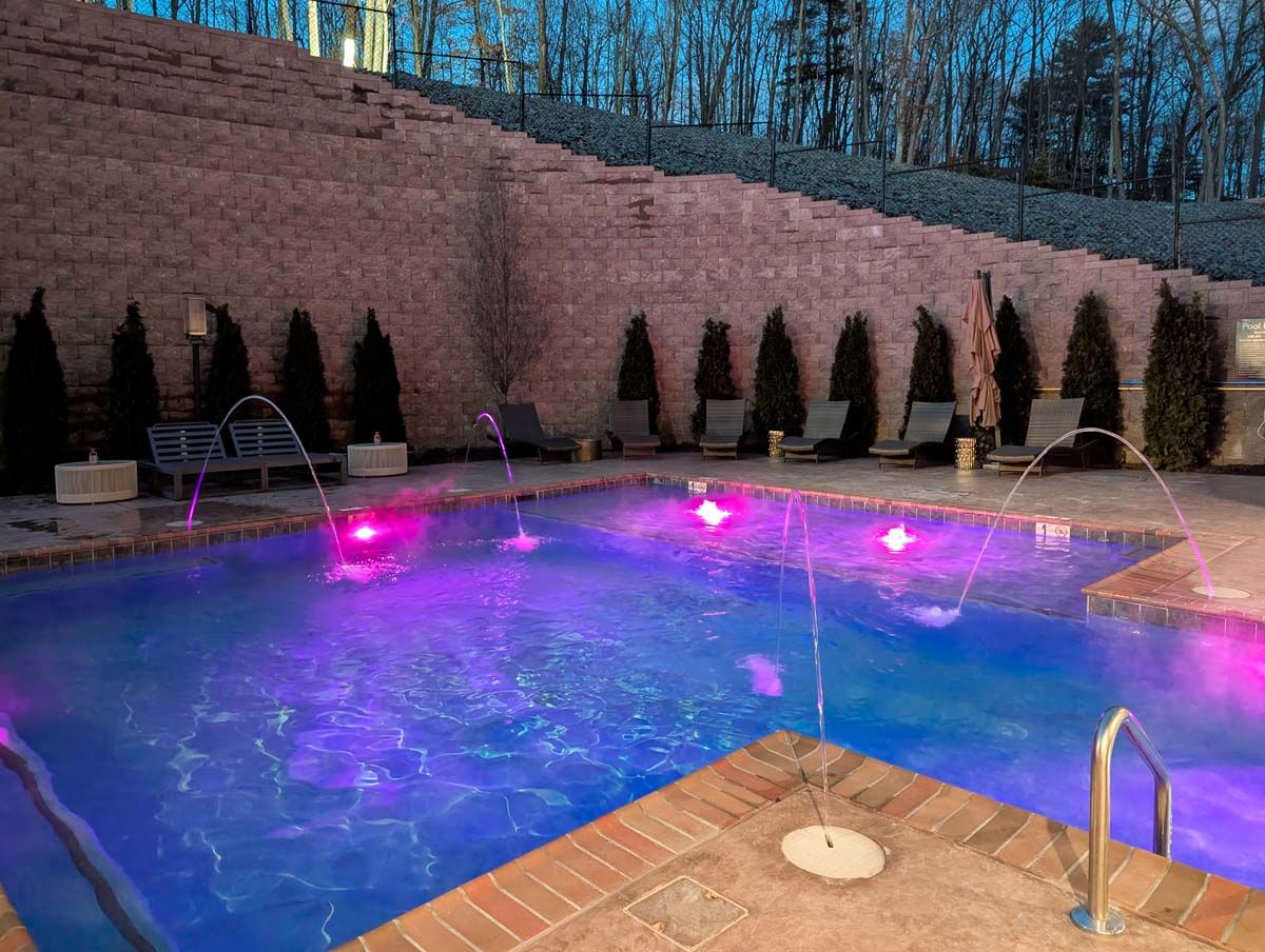 Outdoor pool area illuminated by purple and blue LED lights, featuring arching water features, brick surround, comfortable lounge chairs, and dramatic terraced stone wall with evergreen landscaping