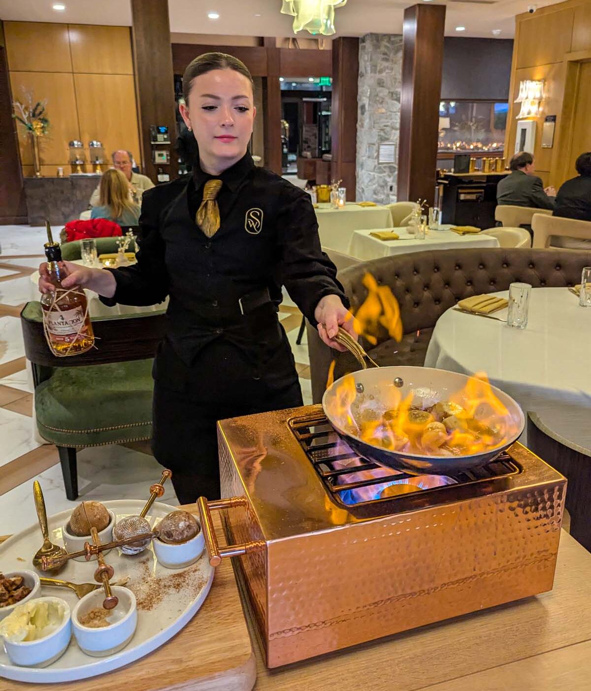 Professional server in black uniform with gold tie preparing Bananas Foster tableside, featuring dramatic flames in copper service pan, surrounded by elegant dining room with crystal chandeliers