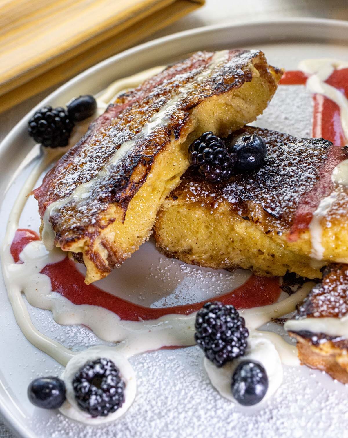 Close-up of caramelized French toast with crispy browned edges, garnished with fresh blackberries and blueberries, dusted with powdered sugar, served over strawberry coulis and crème anglaise